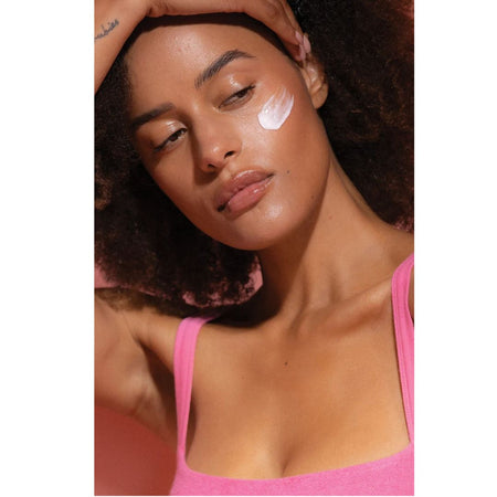 A close-up lifestyle shot of a woman with dark curly hair wearing a bright pink tank top, with a white cream product swatch applied to her cheek.