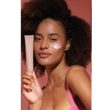 A woman with dark curly hair holds a light pink cosmetic tube next to her cheek, which has a swipe of white cream. She is wearing a pink top against a solid pink background.