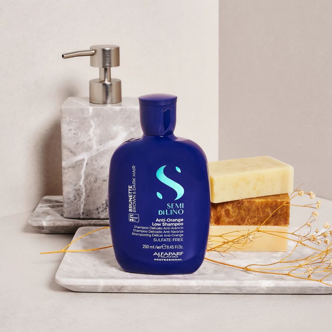 A bottle of Semi Di Lino Anti-Orange shampoo stands on a white marble tray, styled with a soap dispenser, stacked bars of soap, and dried yellow flowers against a neutral beige background.