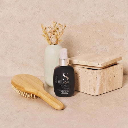 A black hair spray bottle with a silver pump and white text, styled on a beige stone surface next to a wooden hairbrush, a light green ceramic vase with dried grass, and a beige stone box.