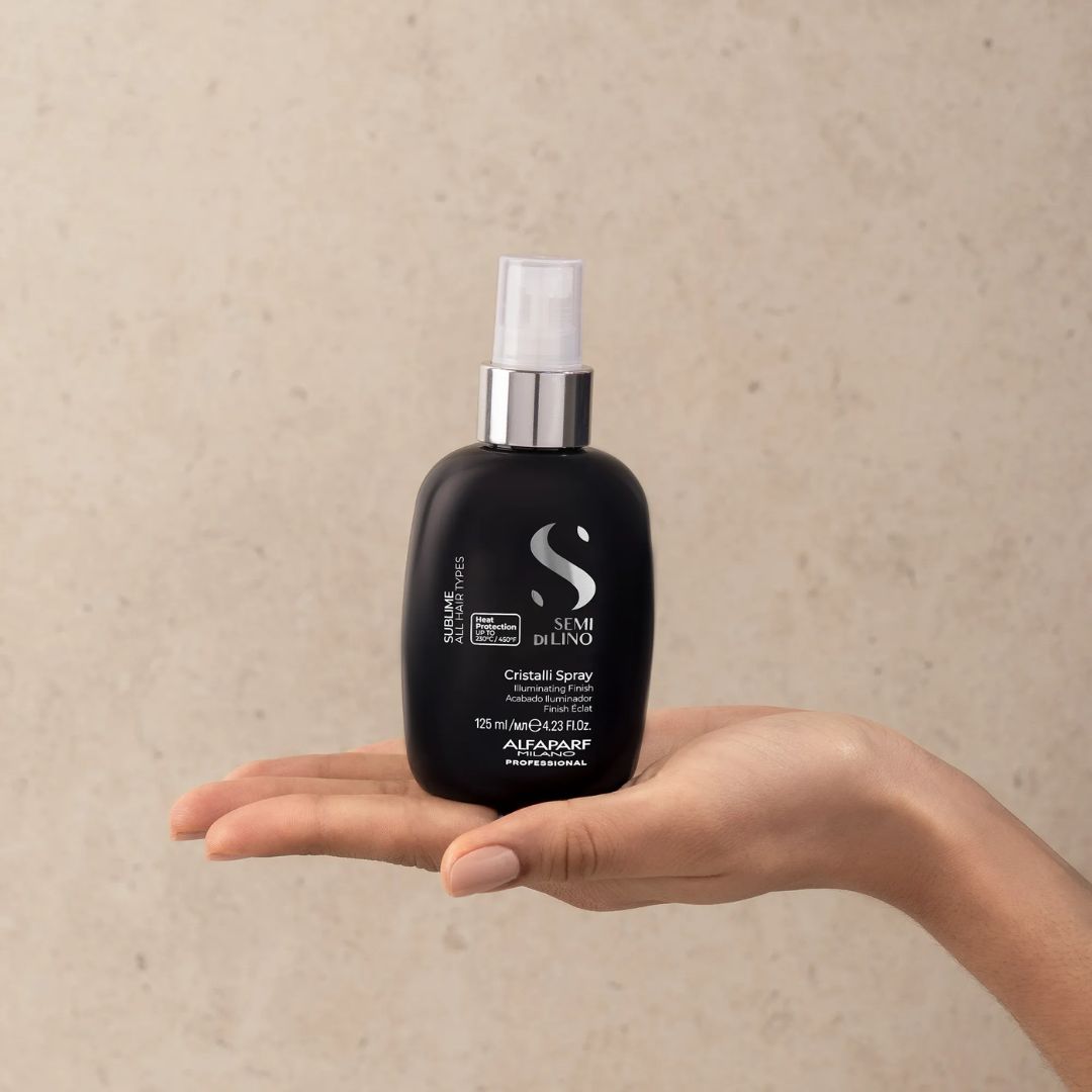 A hand holding a black hair spray bottle with a silver pump and white cap against a textured beige background. The bottle features the Semi Di Lino brand logo and text.