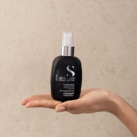 A hand holding a black hair spray bottle with a silver pump and white cap against a textured beige background. The bottle features the Semi Di Lino brand logo and text.