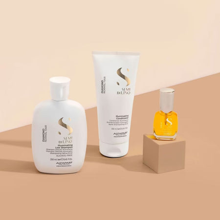 Three  Alfaparf Milano Semi Di Lino Diamond Gift Set haircare products arranged on a beige and peach background: a white shampoo bottle, a white conditioner tube, and a small amber perfume-style oil bottle on a beige pedestal.