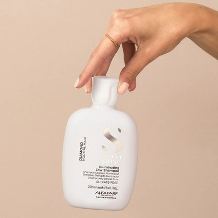 A hand holding a white bottle of Alfairp Professional Diamond Normal Hair Shampoo against a solid beige background.
