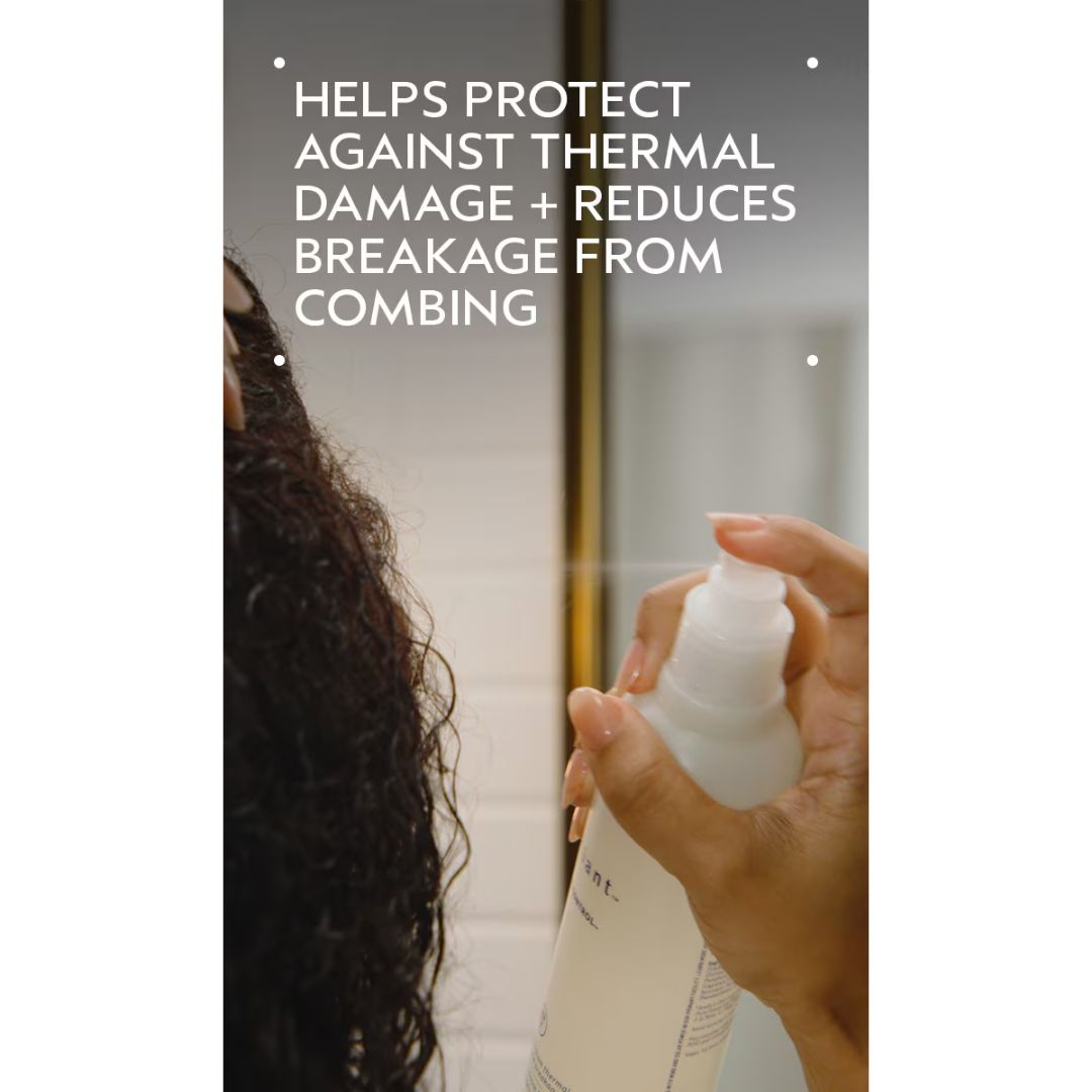 A person with dark curly hair sprays a white pump bottle onto their hair in a bathroom setting. White text at the top reads, 'HELPS PROTECT AGAINST THERMAL DAMAGE + REDUCES BREAKAGE FROM COMBING.