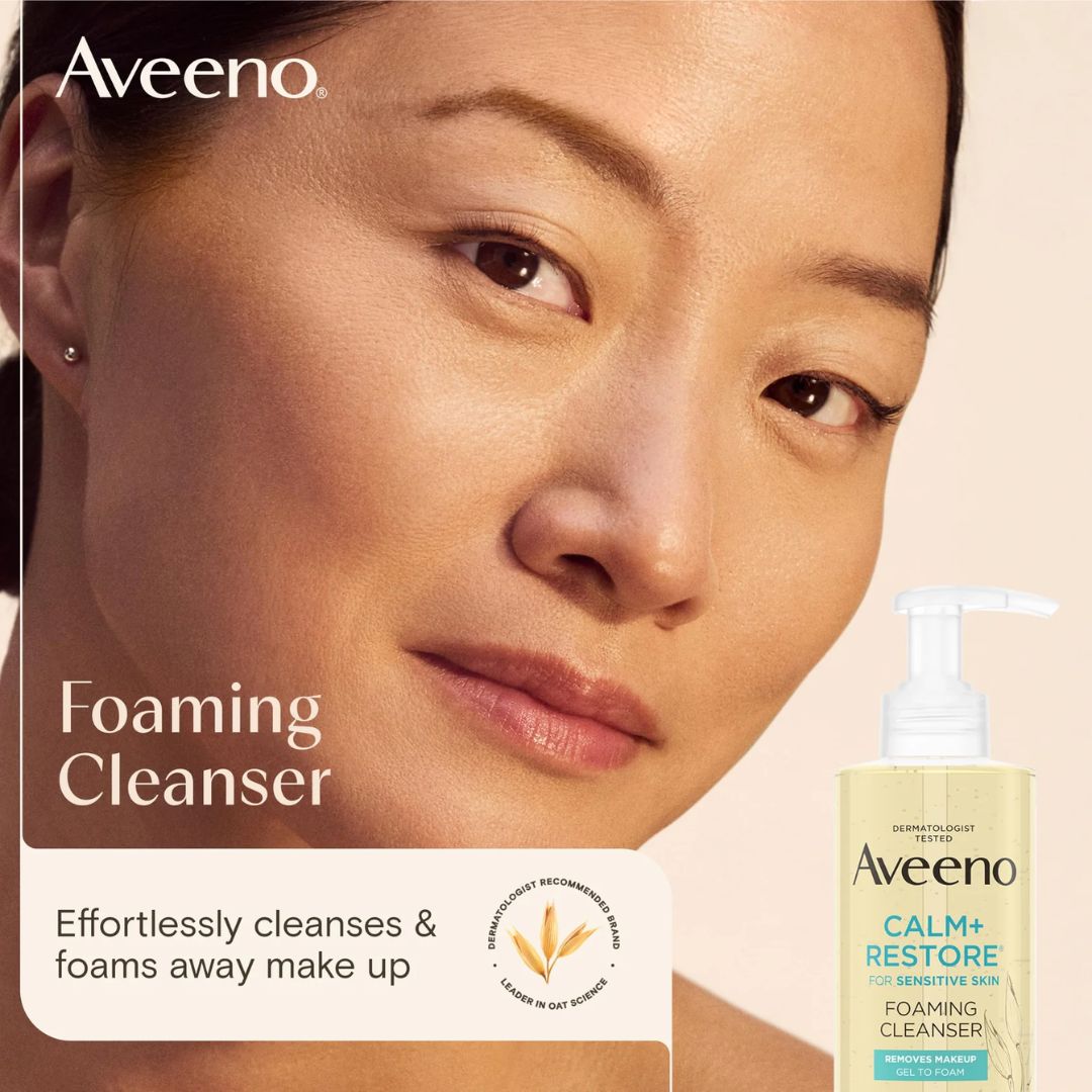 Aveeno Calm+ Restore Foaming Cleanser pump bottle shown next to a close-up of a woman's face with clear, dewy skin. The image features the Aveeno brand logo, product name, and a tagline about cleansing and foaming away makeup.