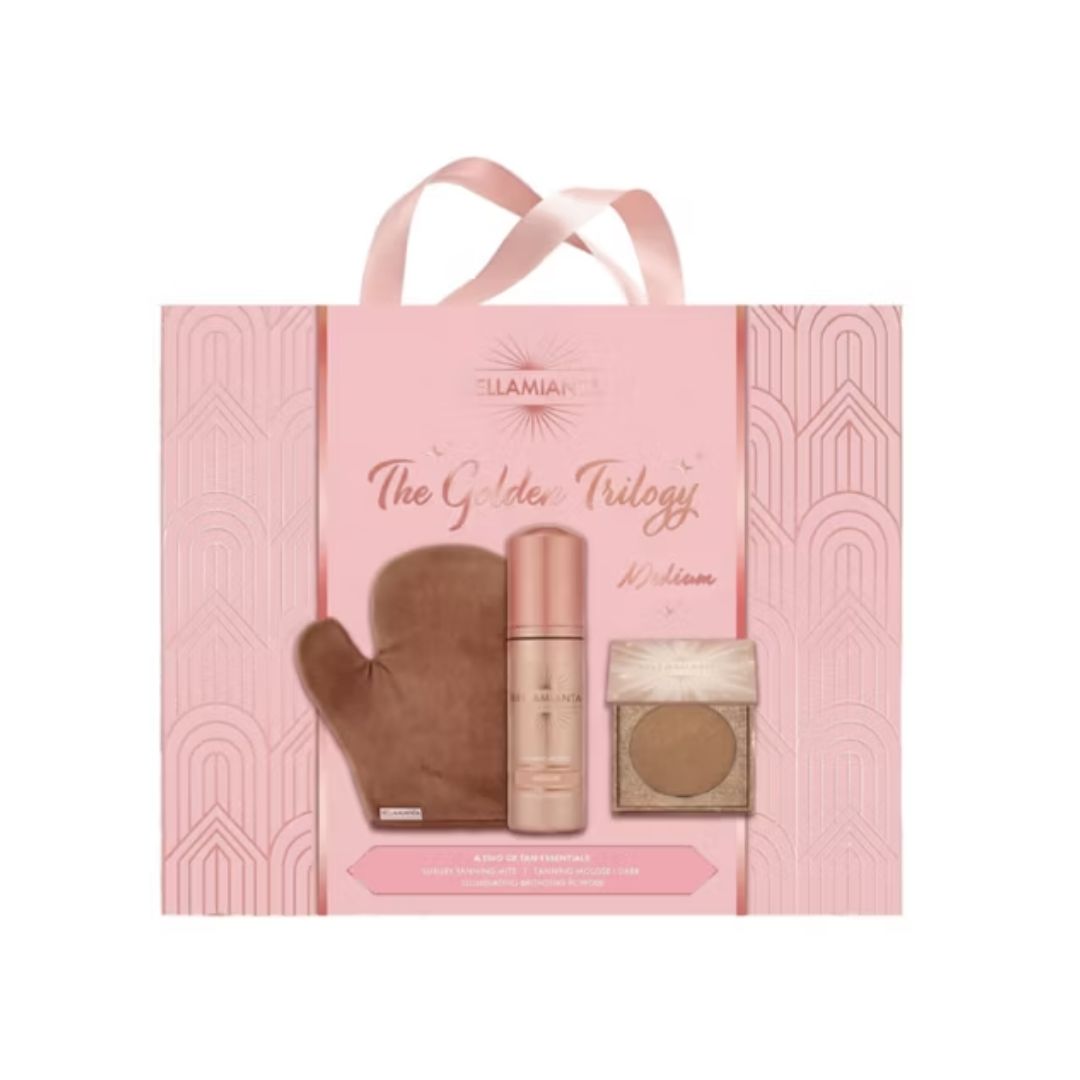 A pink gift bag set from Bellamiantas 'The Golden Trilogy' collection, featuring a brown velvet glove, a metallic gold-toned bottle, and a small square compact with a warm brown powder, all displayed on a clean white background.