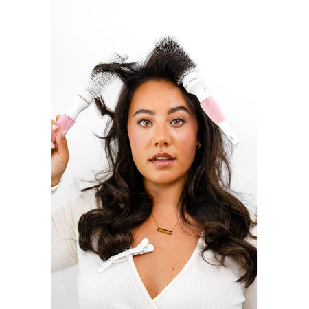 A woman with long dark wavy hair uses a white and pink round hairbrush to style her hair against a plain white background.