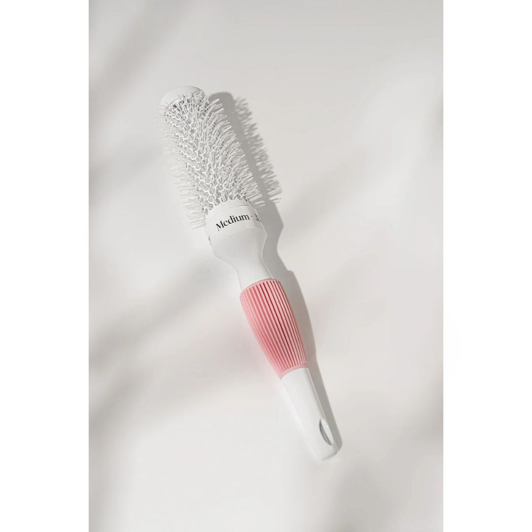 A medium-sized round hairbrush with white bristles, a white plastic head, and a textured pink handle, shown on a white background with soft leaf shadows.