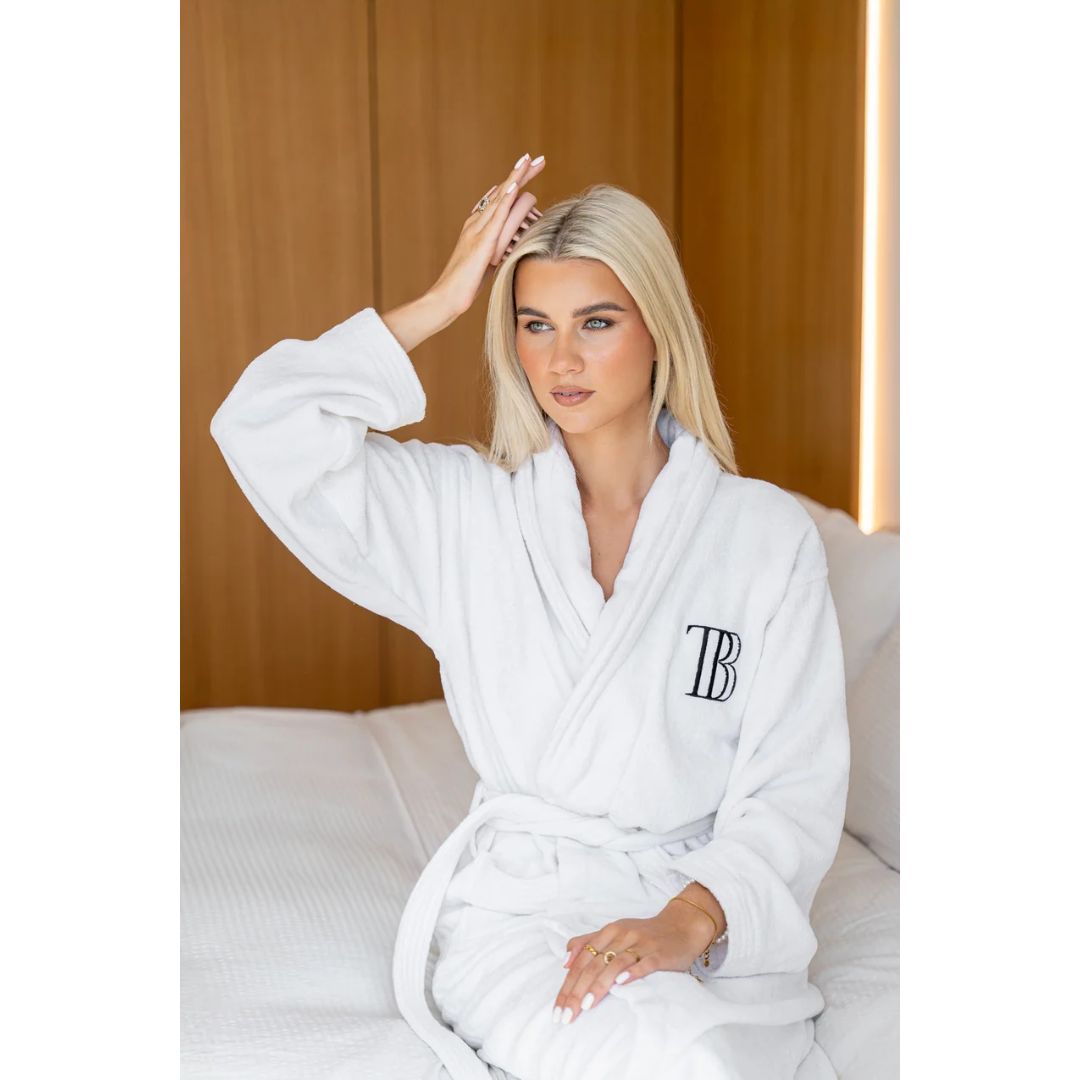 A blonde woman sits on a bed wearing a white plush bathrobe with a black embroidered monogram 'TB' on the left chest. The background features a warm wood-paneled wall.