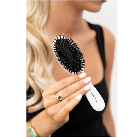 A close-up of a woman holding a white oval hairbrush with black bristles. The brush has the text 'The Hairbrush' printed on the handle.