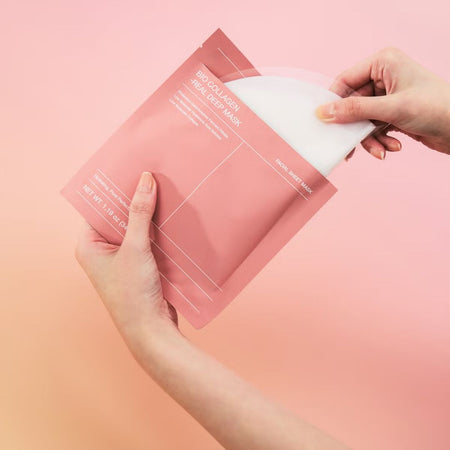 A person's hands are shown peeling back a white sheet from a pink resealable pouch of a facial sheet mask, set against a solid pink background.