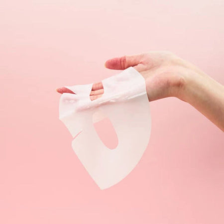 A hand holding a translucent white sheet mask against a solid light pink background.
