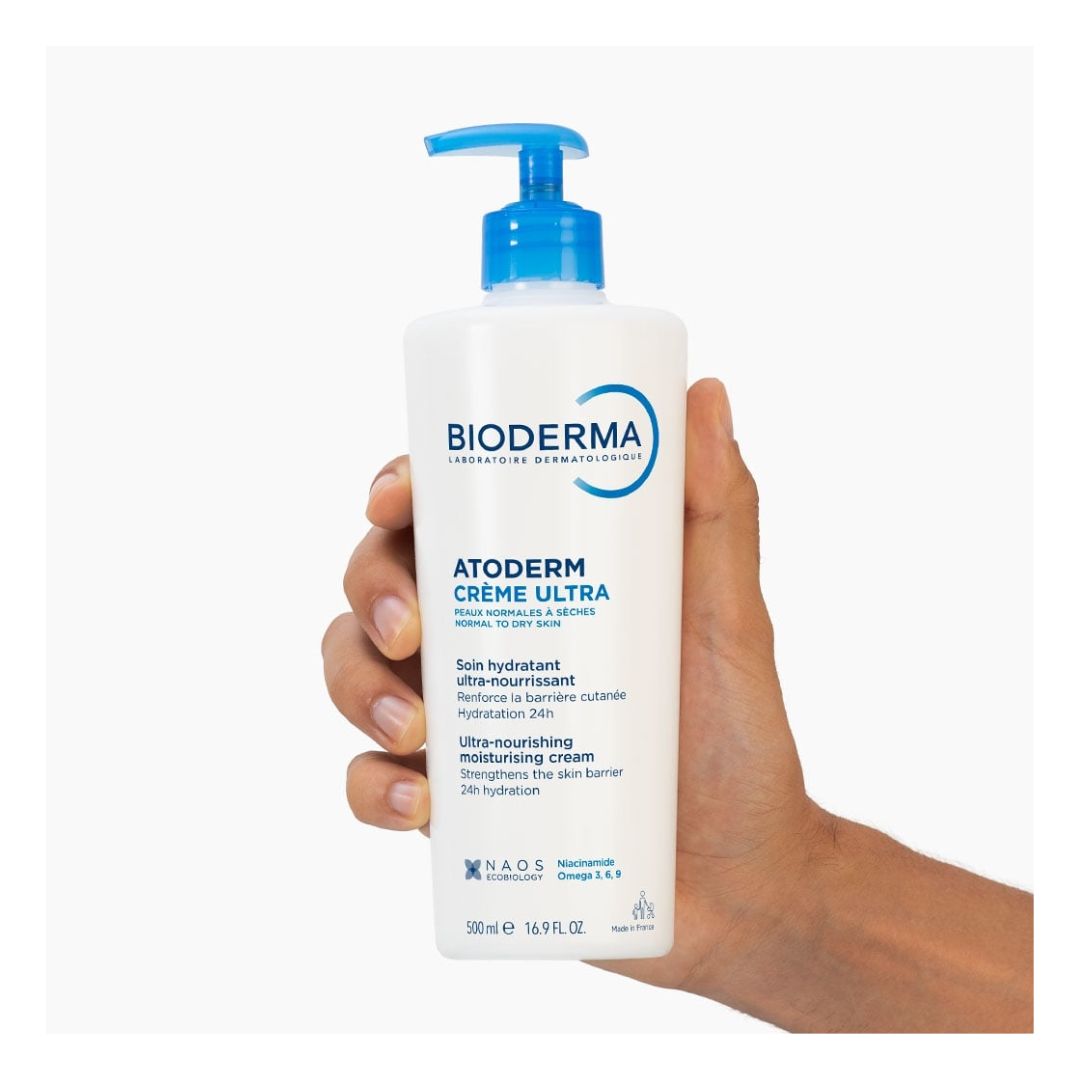 A hand holding a 500ml white bottle of Bioderma Atoderm Crème Ultra moisturizing cream against a plain white background.
