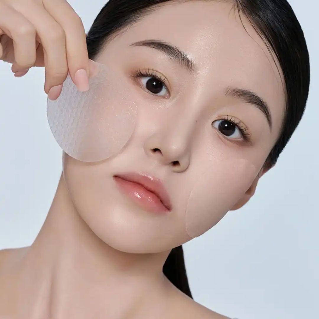 A close-up of a woman holding a translucent circular patch against her cheek, with a second patch applied to her forehead and temple. She has a clean, glossy complexion against a light blue background.