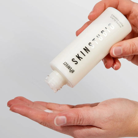 Hand holding a bottle of SKIN STUDIO product with a small amount on another hand against a light gray background
