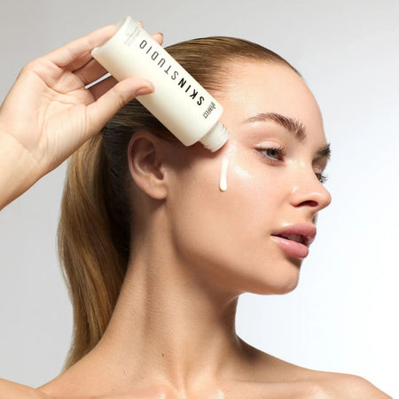 Woman applying skincare product to her face with a focus on the brand 'SKINSTUDIO'.
