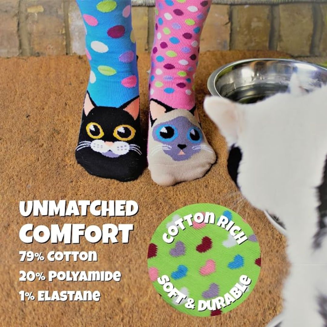 A pair of mismatched cat-themed socks worn on a person's feet, with a real cat partially visible on the right and a bowl of water nearby. Text on the image highlights the socks' comfort and material: 79% cotton, 20% polyamide, 1% elastane.