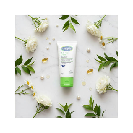 A tube of Cetaphil Moisturizing Cream centered on a white marble flat-lay background, surrounded by white roses, daisies, green leaves, and small pearl-like beads.