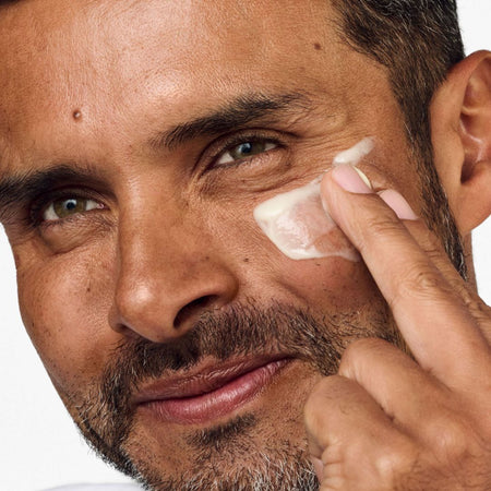 Close-up of a man applying a white cream or balm to his cheek with his finger against a plain light background.
