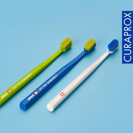 Three Curaprox toothbrushes in green, blue, and white on a light blue background.