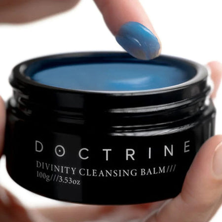 Person applying blue cleansing balm from a black jar labeled 'Doctrine' on a white background