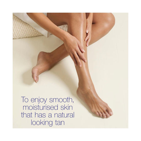 A person sitting on a cream-colored floor or cushion, with their legs crossed and hands resting on their shin. Blue text at the bottom reads, 'To enjoy smooth, moisturised skin that has a natural looking tan'.