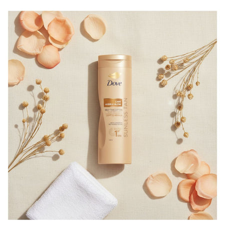 A bottle of Dove Sunless Tan Care Deep Tan body tan lotion is centered on a textured beige background, styled with peach rose petals, dried floral sprigs, and a folded white towel.