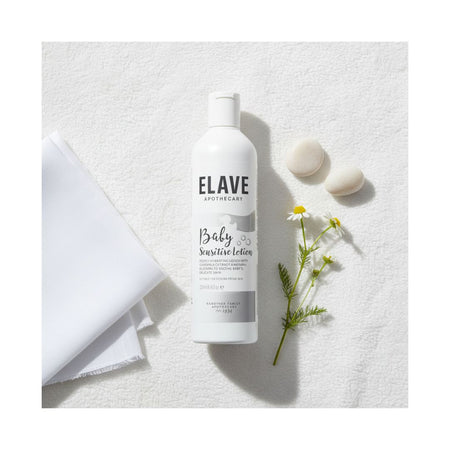 A bottle of ELAVE APOTHECARY Baby Sensitive Lotion is displayed in a styled flat-lay arrangement on a textured white surface, accompanied by a folded white towel, two smooth stones, and a small sprig of chamomile flowers.