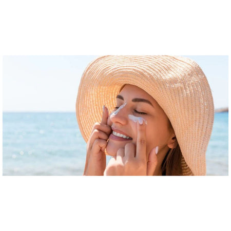 A woman wearing a large woven straw sun hat applies sunscreen to her face with her fingers, set against a blurred beach background with blue water and sky.