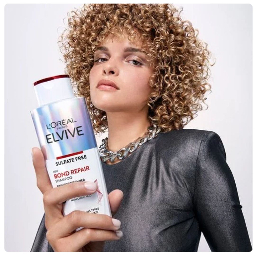 A woman with voluminous curly hair holds a bottle of L'Oréal Paris Elvive Bond Repair Shampoo. She is wearing a dark metallic gray top and a chunky silver chain necklace against a clean, light gray studio background.