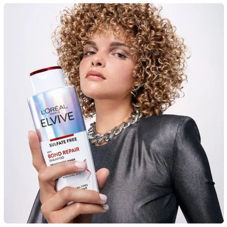 A woman with voluminous curly hair holds a bottle of L'Oréal Paris Elvive Bond Repair Shampoo. She is wearing a dark metallic gray top and a chunky silver chain necklace against a clean, light gray studio background.