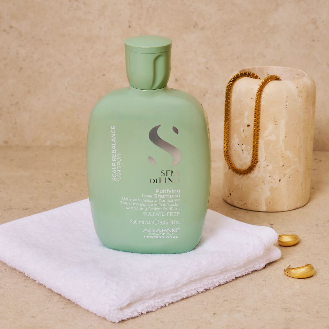 A bottle of Alfarab Sen DiLin Scalp Balance shampoo in a sage green color, styled on a white towel next to a stone cylinder with a gold chain.