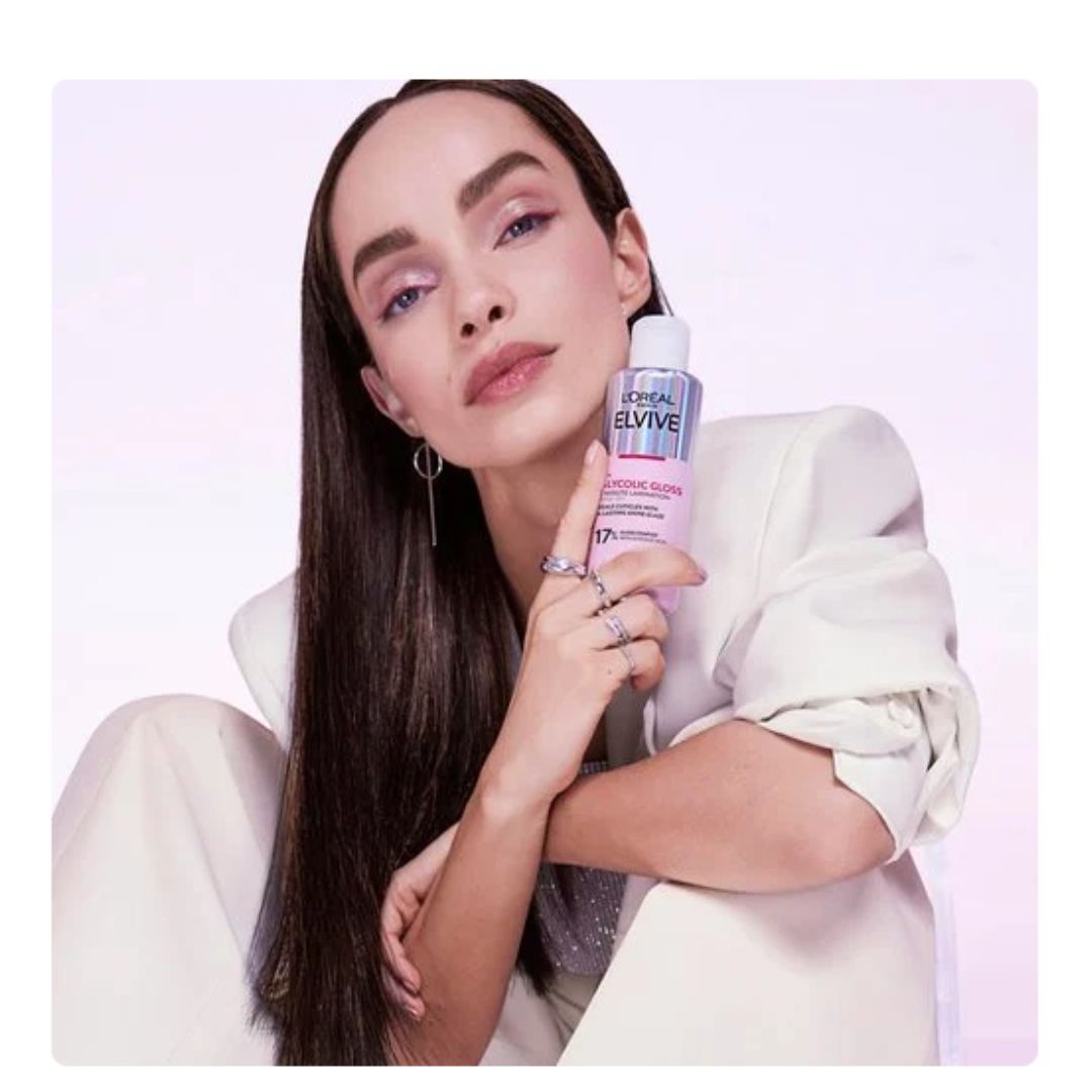 A woman with long dark hair wearing a cream blazer holds a bottle of L'Oréal Paris Bluvite Hair Color Remover against a light pink studio background.
