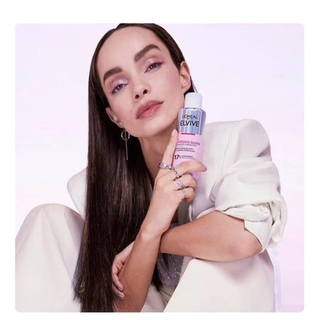 A woman with long dark hair wearing a cream blazer holds a bottle of L'Oréal Paris Bluvite Hair Color Remover against a light pink studio background.