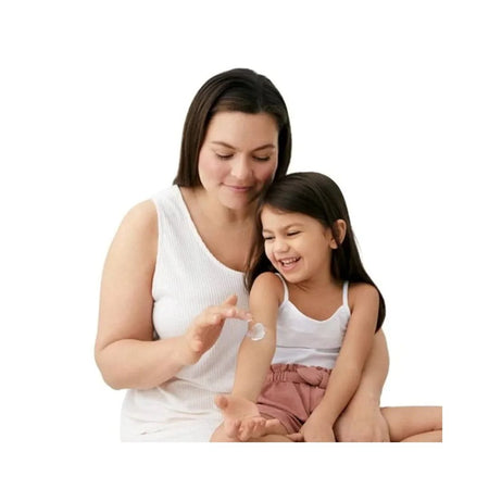A woman and a young girl sitting together, applying white cream from a small glass bottle onto the girl's arm against a plain white background.