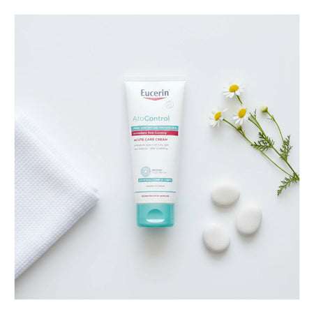 A tube of Eucerin AtoControl Active Care Cream is displayed in a flat-lay composition on a white background, accompanied by a folded white textured towel, three white stones, and a sprig of white chamomile flowers.