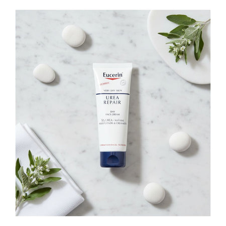 A tube of Eucerin Urea Repair Day Face Cream is displayed on a light marble surface, styled with white soap bubbles, green leaves, and folded white towels.