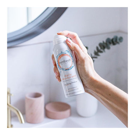 A hand holding a can of Memfresh Daily Deodorant in a bright bathroom setting with a sink, mirror, and small decorative items in the background.