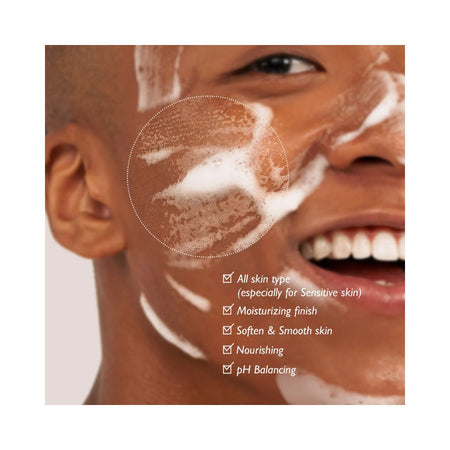 Close-up of a smiling man's face with white foaming cleanser on his cheek, overlaid with a dotted circle highlighting the cleanser texture and a list of benefits including all skin type, moisturizing finish, soft & smooth skin, nourishing, and pH balancing.