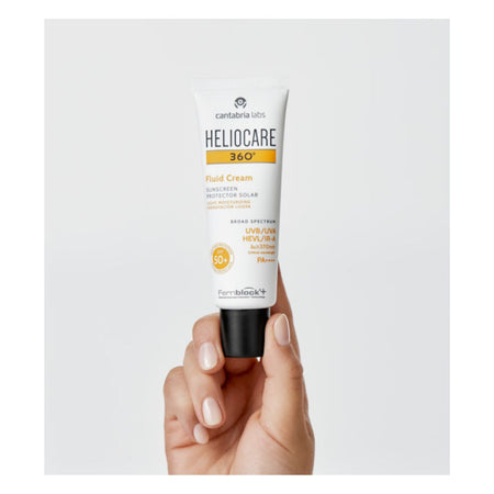 A hand holding a white tube of Caratina Bio Heliocare 360° Fluid Cream sunscreen against a light gray background.