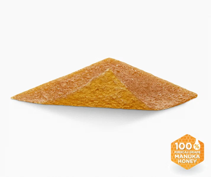 A triangular pyramid-shaped piece of golden-brown Manuka honey, shown on a clean white background with a small orange '100% Pure Manuka Honey' badge in the bottom right corner.