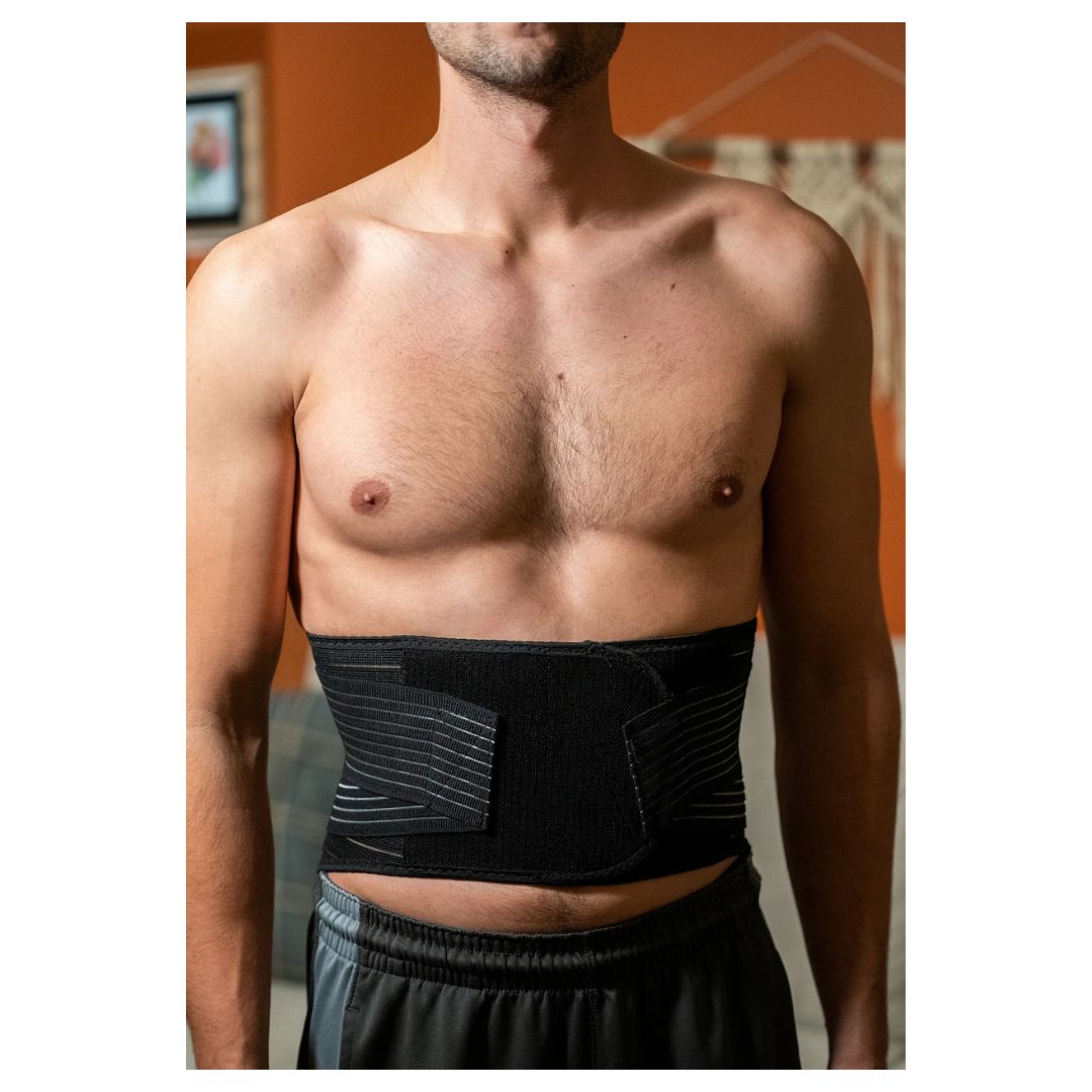 A man wearing a black abdominal support belt with textured side panels, shown from the chest down in an indoor setting.