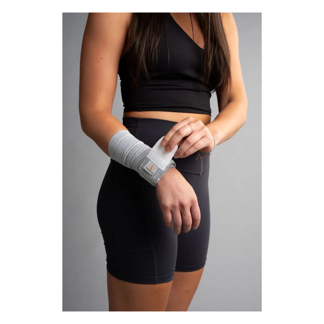 A woman wearing a black sports bra and black athletic shorts is shown from the chest down, demonstrating a white wrist wrap with a clear plastic fastener on her left wrist, against a solid gray studio background.