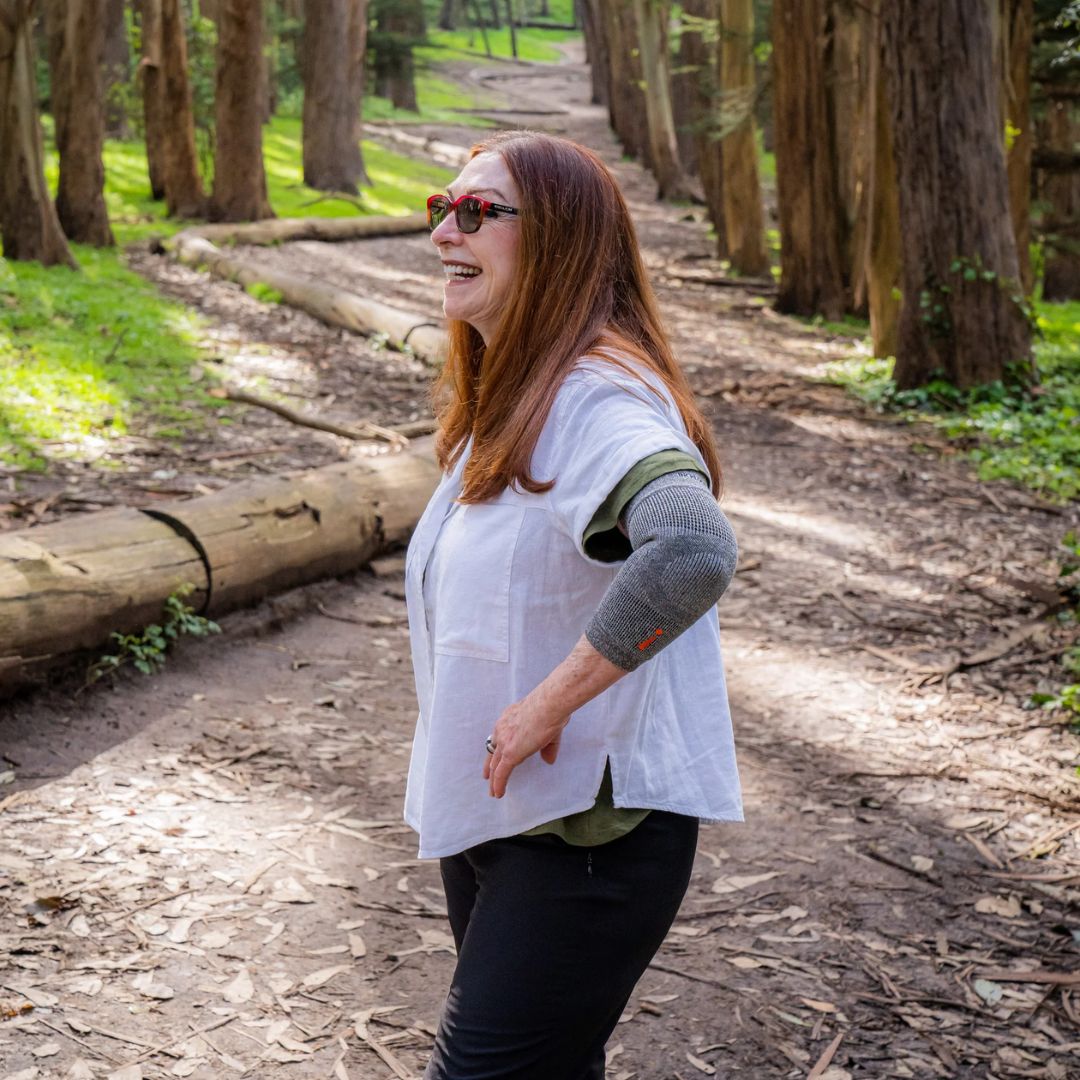 A woman wearing a white sleeveless vest over an olive green shirt and a gray knit elbow sleeve, standing on a dirt path in a wooded outdoor setting.