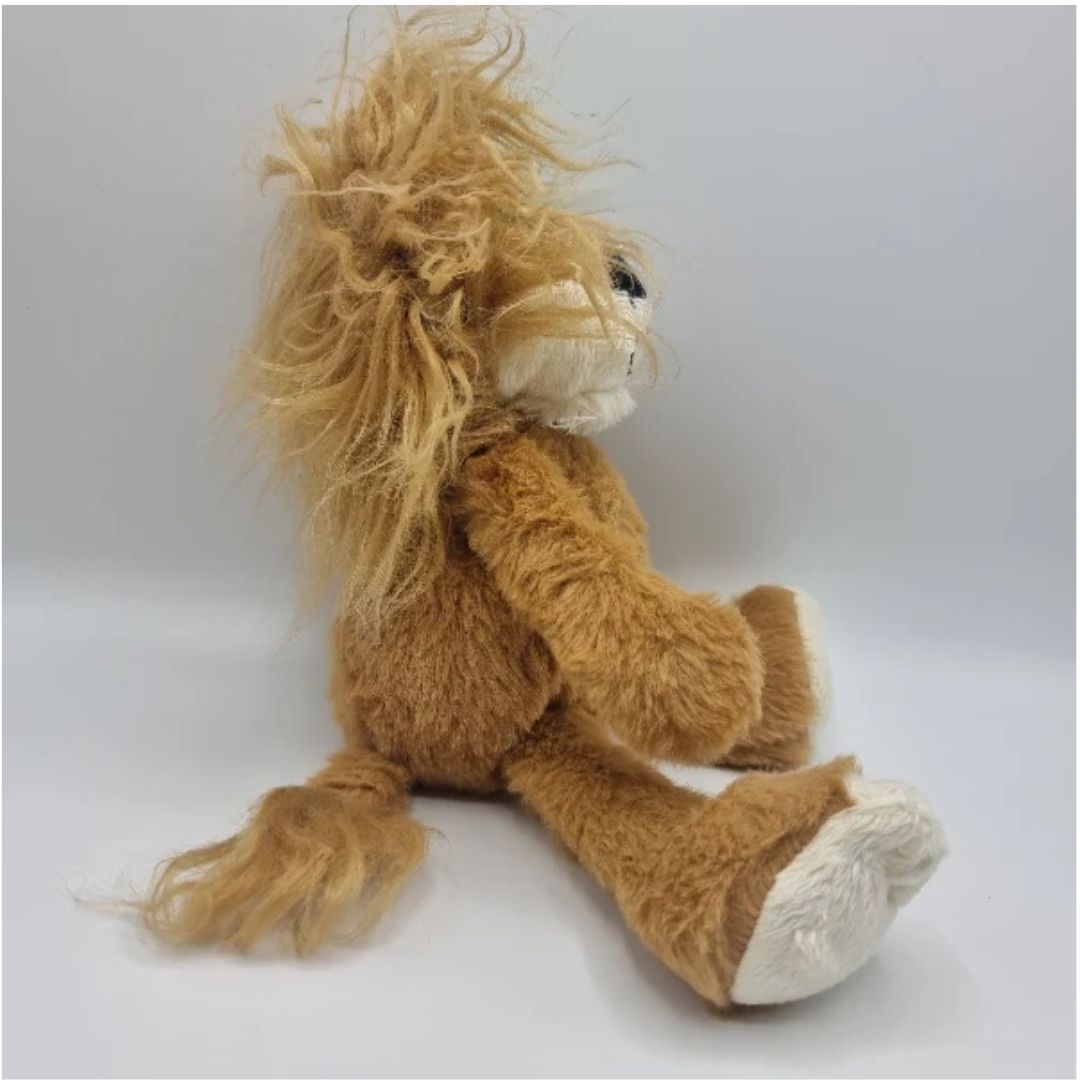 A plush lion toy sitting on a light gray studio background, with a tan-brown body, long shaggy golden-brown mane, white muzzle and feet, and small black eyes.