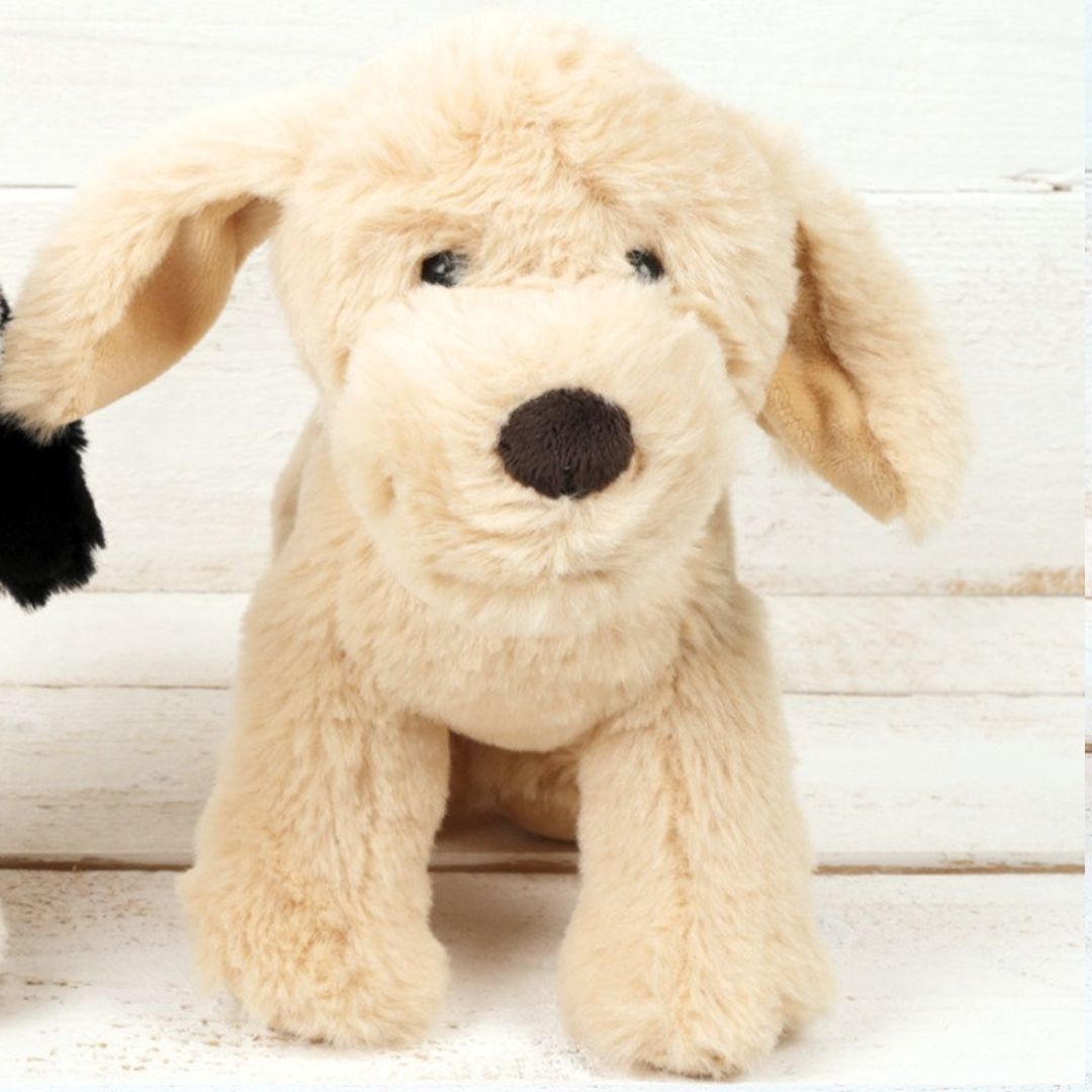 A plush puppy toy with cream-colored faux fur, floppy ears, a dark brown nose, and black eyes, sitting against a white wooden plank background.