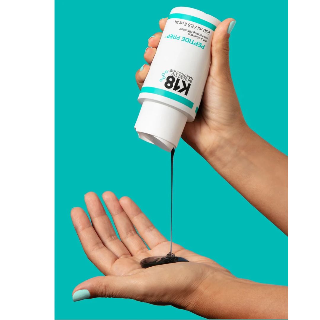 A hand holds a white tube of K18 Peptide Pimp serum, pouring a dark black gel into the palm of another hand against a solid teal background.