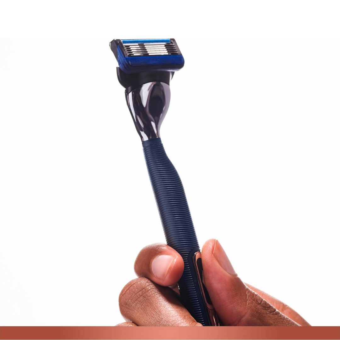 A hand holding a four-blade manual razor with a dark blue textured handle, metallic silver neck, and blue and black head against a white background.