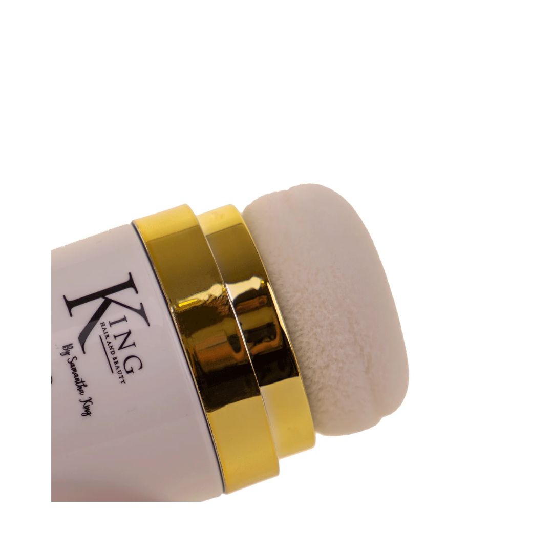 Close-up of a white cylindrical cosmetic container with shiny gold metallic bands and a soft off-white sponge applicator, on a plain white background. The container shows the brand text 'KING' 
